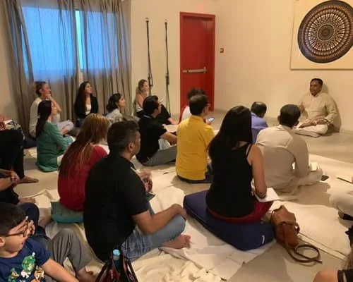 A group seated on the floor in a quiet room, listening to a teacher at a session